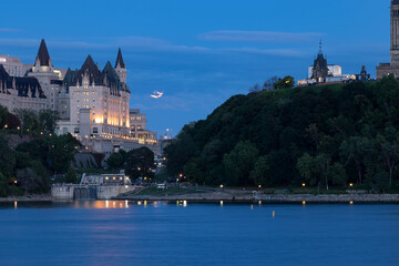 Historic Chateau Laurier and downtown Ottawa at night from across the Rideau River.  Blue night sky with a full moon partially covered by a cloud completes the fairytale view.