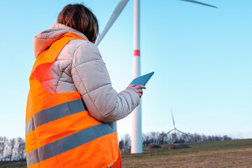 A woman engineer, holding a tablet and working at the wind turbine farm in the field in the orange...