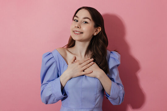Thankful, grateful young woman keeps hands on cheast, breast looking at camera, pink background, shadow. Happy smiling girl in casual shirt, natural makeup studio shot. Cheerful expression