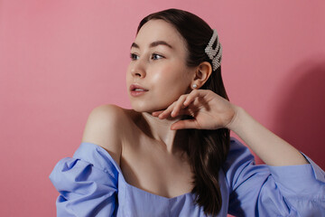 Young woman in blue shirt with naked shoulders, natural makeup and curly hairstyle looking at camera over pink background, shadow. Girl in casual outfit in pearl earrings and hairpins studio shot.