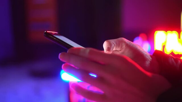 A woman holds a phone in her hands at night or in the evening. It is illuminated by blue and pink neon light. Club or casino lighting, advertising sign. It's cold outside, warm clothes, close-up.