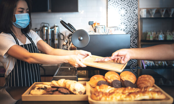 Asian woman, Coffee shop business owner wearing a surgical mask, accept cash payments from customers, avoiding contact By placing money on a wooden tray, to preventing the spread of virus concept. - Powered by Adobe