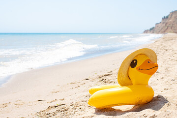 Fototapeta na wymiar Yellow inflatable duck ring in summer straw hat laying on sandy empty beach near blue wavy ocean in sunny day. Protection swim tube for kid. Summer travel vacation resort concept. Copy space