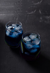 Two glasses with a blue cold drink with pieces of ice on a black background in a dark key, vertical background, selectiv focus