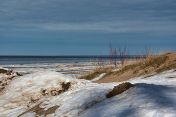 The landscape of the coastal strip of the Baltic Sea in early spring, the last snow and sparse vegetation on the sand against the background of the sea with grayish ice and cloudy sky.