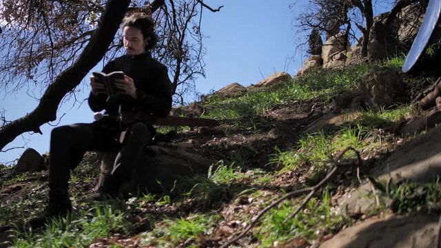 Man in medieval outfit sits on rock and reads a book. 4k, 60fps, tracking shot