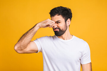 Caucasian bearded man isolated over yellow background smelling something stinky and disgusting, intolerable smell, holding breath with fingers on nose. Bad smells concept.