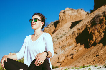 Fototapeta na wymiar Happy young woman practicing breathing yoga, sitting in lotus pose on sandy brown cliff and blue sky background. Smiling female athlete meditating outdoor in sunny day,enjoying outdoor training class