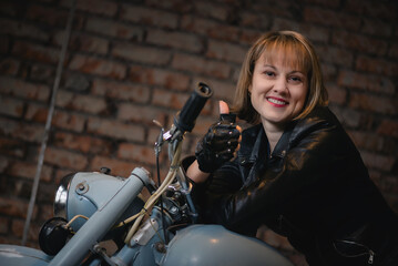 Fototapeta na wymiar Happy young motorbiker woman is sitting on the retro motorbike and shows a thumbs up gesture in the old garage.