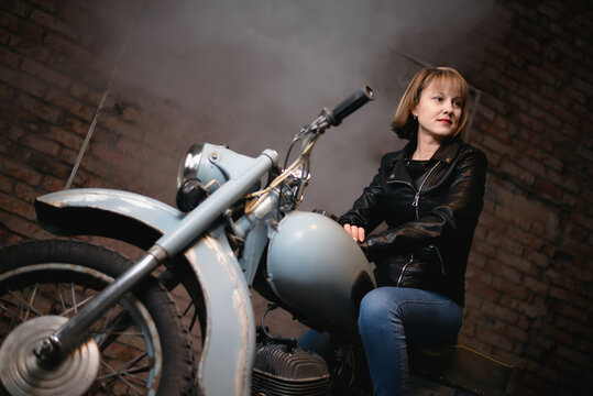 Young biker woman is sitting on the retro motorbike in the old garage.