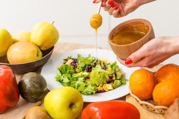 Woman pouring honey mustard dressing into bowl with fresh salad on table