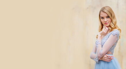 Portrait of a young blonde woman in a delicate blue dress against a beige wall