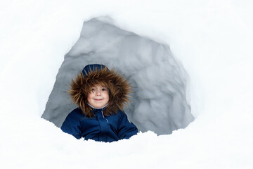 Chilld in a winter jacket with a fur collar sits inside an ice house, an igloo, a shelter. Winter game, playing Eskimo. - obrazy, fototapety, plakaty