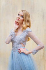 Portrait of a young blonde woman in a delicate blue dress against a beige wall. Blonde girl with beautiful styling