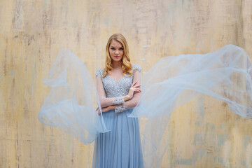 Portrait of a young blonde woman in a delicate blue dress against a beige wall. The dress is semi-transparent for the bride's morning, it develops