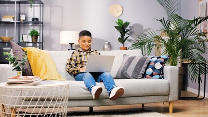 Cheerful cute little African American boy child sitting on sofa in cozy living room at home and browsing online on laptop watching cartoons and smiling, happy childhood, kid concept,