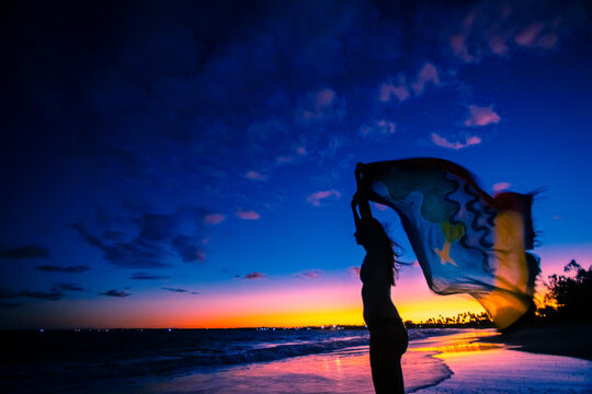 Silhueta de mulher com os bra&ccedil;os levantados, segurando uma sa&iacute;da de banho, olhando o horizonte durante p&ocirc;r-do-sol na praia de Serrambi.
