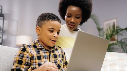 Close up of African American joyful young small cute son browsing online on laptop sitting on sofa with beautiful mother. Little boy with mom studying on computer surfing internet, childhood concept
