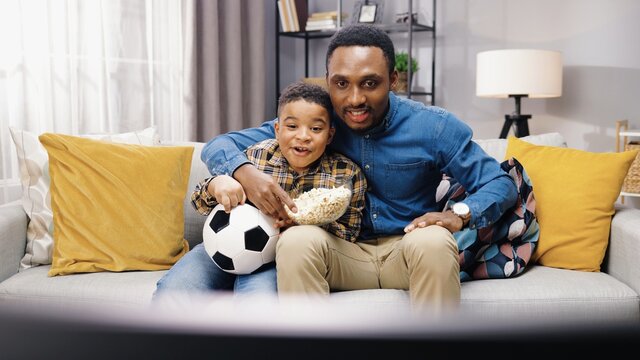 Portrait Of African American Cheerful Young Father Spending Time With Small Son Sitting On Couch In Apartment And Eating Popcorn While Cheering For Favorite Football Team Watching Game Match On TV