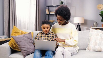 Portrait of happy African American family pretty mother and small son child sitting on sofa browsing online on laptop at home. Mom and little boy studying on computer, e-learning, education concept