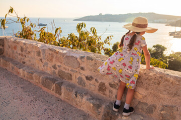 A little girl with a big hat looks at the sea and the city. A girl in the castle of Bodrum, Turkey. The baby is resting and going to the sights.