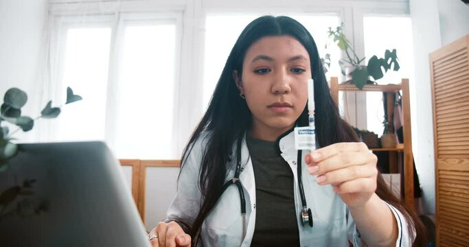 Zoom in on professional multiethnic doctor woman in lab coat looking at new vaccine syringe cure, checking with laptop.
