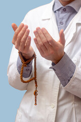 A young man in a white robe raised his hands up and prayed with a Muslim rosary. Blue background. Doctor praying. A Muslim makes dua