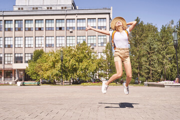 Obraz premium Happy young woman celebrate end of lockdown. Borders are open. Jumping at street if town. Summer time. Staycation concept. Female tourist. Backpack, hat, t-shirt, shorts. Sunny day