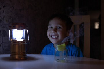 a child in a blue T-shirt is sitting at a table, smiling, and there is a flashlight and an...