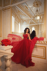 Beautiful brunette girl in a red ball gown on the background of white and gold palace interiors