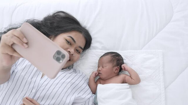 Mother and her newborn baby making a selfie or video call to family on the bed white in home. Mixed race black boy Ethnicity Thai-Nigeria. New generation Technology Communication