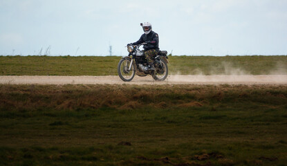 a motorcyclist rides his classic Royal Enfield four stroke motorbike along a dusty stone track over...