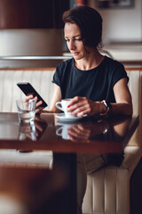A woman in a black T-shirt is sitting alone at the bar, using the phone and drinking coffee