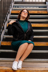 Fototapeta na wymiar Stylish young pretty hipster woman sitting on iron house stairs, Happy beautiful girl in casual clothes green dress, black jacket white sneakers, urban background, street photo, body positivity