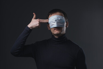 Adult man with medical mask on his eyes showing gesture of shooting himself. Concept of anxiety and panic during global lockdown.