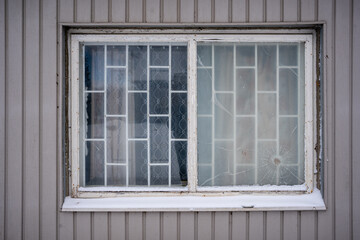 An old window with white grilles is broken and cracks have formed in it, which form an interesting pattern, but the facade is lined with gray boards vertically.
