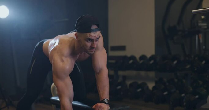 Handsome shirtless sporty muscular bodybuilder man excercise with weights building his back muscle in professional sport gym. Bodybuilder lifts dumbbell on bench, slow motion