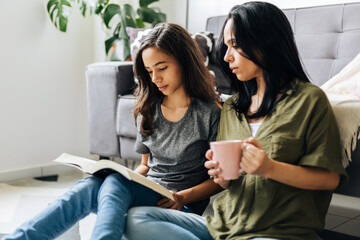 Single parenthood. Mother and daughter reading a book together at home. - obrazy, fototapety, plakaty