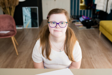 Naklejka na ściany i meble Portrait of cute teenager girl with Down syndrome sitting at home and looking at camera. Domestic life of people with disabilities. World Down syndrome day.
