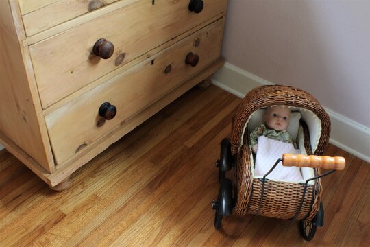 Small woven baby carriage with fake baby inside a play room with wooden floors and cabinets. 