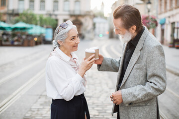 Side view of smiling aged couple in stylish outfits toasting with carton cups of hot coffee while standing on street. Happy mature family smiling and looking kindly on each other.