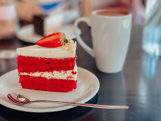 Piece of cake in white and red colors with sponge cake, cream and decoration on top of the cake in the form of strawberry on the table in the restaurant.