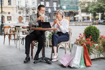 Elderly loving couple sitting at cafe terrace with tablet and retro camera on table, drinking coffee and looking at each other. Colorful shopping bags standing near. Enjoyment of retirement.