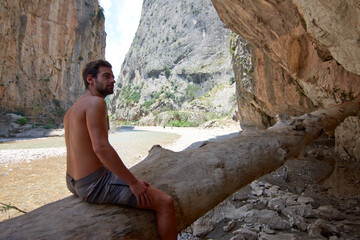 Fototapeta na wymiar A closeup of a half-naked Caucasian man sitting on a big wood log under a stone cave