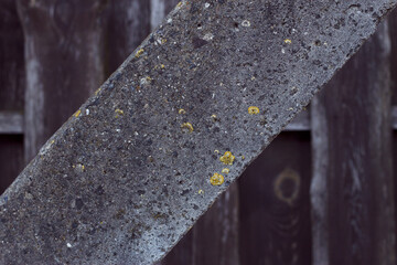 Grey concrete pillar with lichen and old wooden fence on the backdrop. Textured background. Copy space for text on diagonal surface.