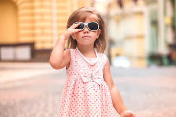 Summer Lifestyle Stylish girl corrects sunglasses. child walks in a European city street. Fashionable kid