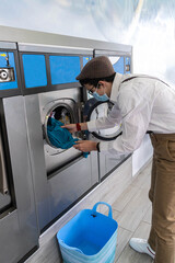 young man in mask white shirt with suspenders taking clothes out of the washing machine after laundry hygiene concept young people