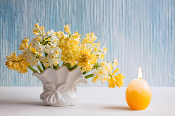 Easter background card with yellow flowers hyacinths, daffodils in a vase and a candle in the shape of an egg on the table.