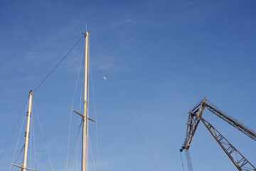 The yacht's masts and an old crane against the blue sky with the moon.