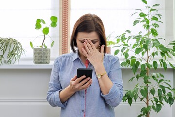 Sad middle aged woman with phone at home near the window